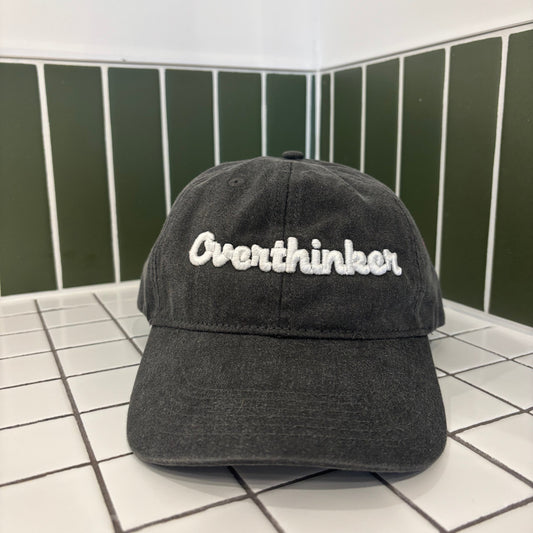 Black cap with 'Overthinker' text on a tiled floor