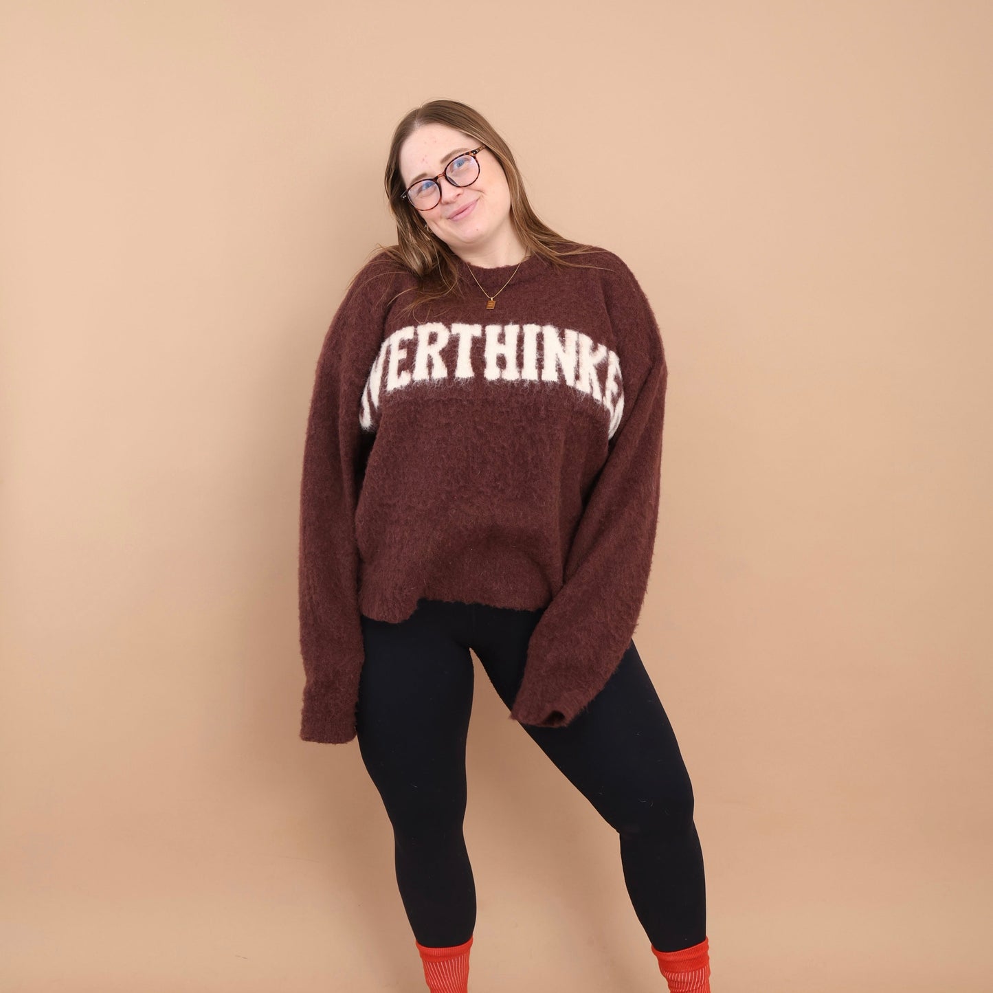 Person wearing a brown sweater with text, black leggings, and red shoes on a beige background