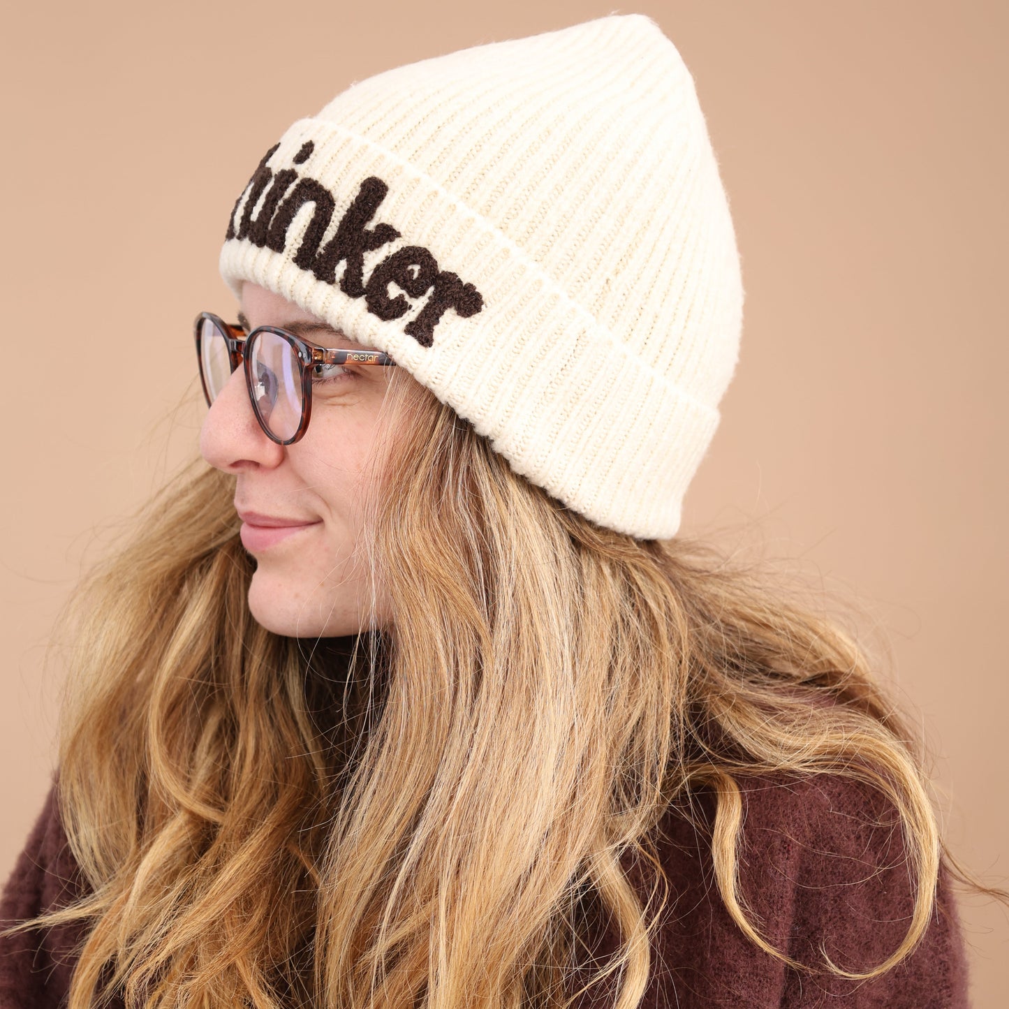 Person wearing a white beanie with text, glasses, and a brown and white patterned garment on a beige background