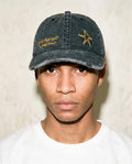 A man wearing a washed black baseball cap with yellow embroidered text and a star logo, paired with a white t-shirt against a textured beige wall.