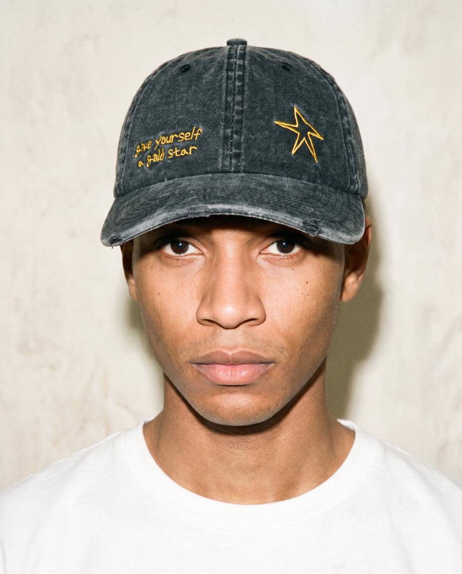A man wearing a washed black baseball cap with yellow embroidered text and a star logo, paired with a white t-shirt against a textured beige wall.