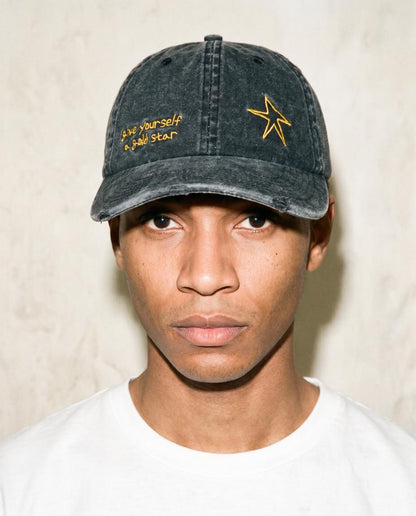 A man wearing a washed black baseball cap with yellow embroidered text and a star logo, paired with a white t-shirt against a textured beige wall.