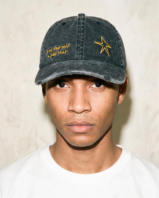 A man wearing a washed black baseball cap with yellow embroidered text and a star logo, paired with a white t-shirt against a textured beige wall.