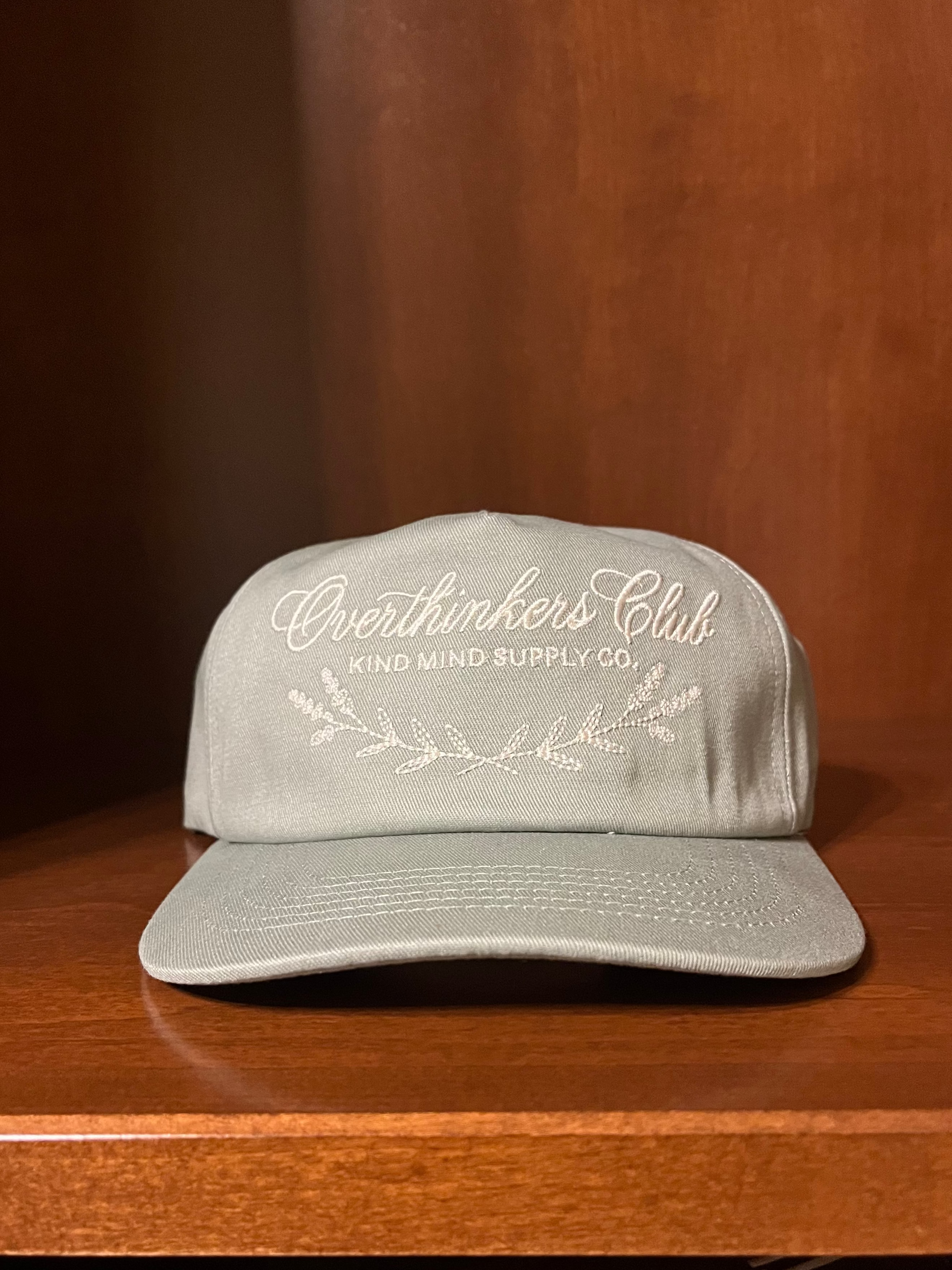A light gray snapback cap with white embroidered text reading 'Overthinkers Club' and 'KIND MIND SUPPLY CO.' on the front, displayed on a wooden shelf.