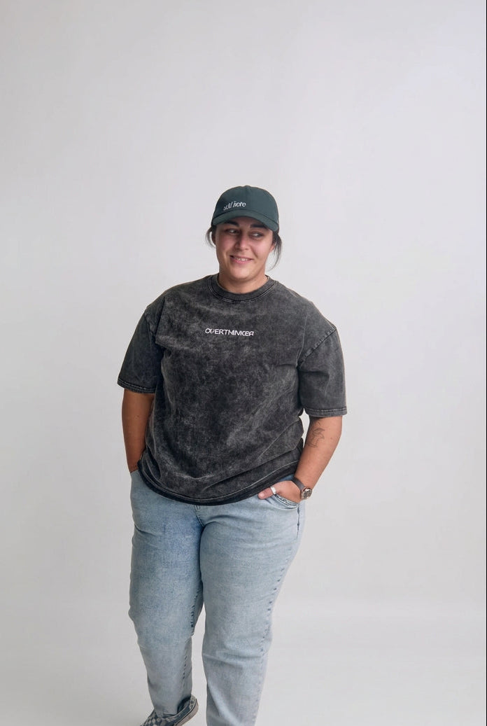 A person standing in a studio wearing a dark gray washed t-shirt with a white logo, light blue jeans, and a dark green baseball cap.