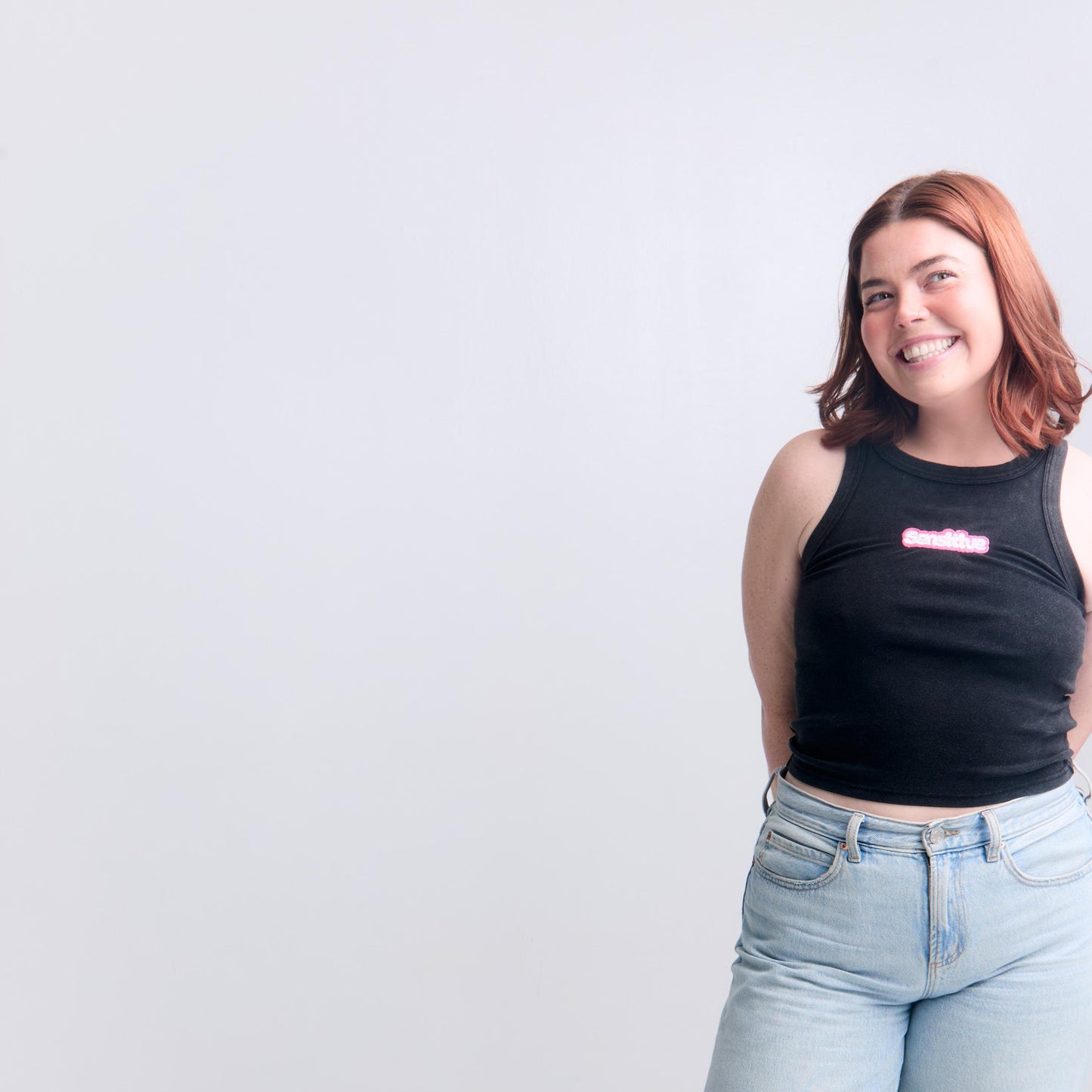 Person wearing a black tank top with pink text and light blue jeans on a white background